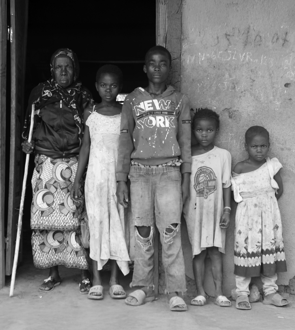 Four serious children standing next to their grandmother in front of a home.